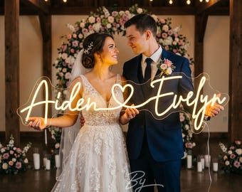 May include: A couple stands in front of a floral arch, holding a neon sign. The sign, in a flowing script, reads "Aiden & Evelyn" with a heart symbol. The bride wears a white wedding dress, and the groom is in a navy suit. The background includes candles and string lights.