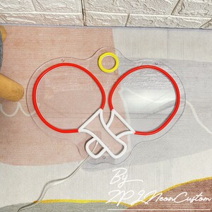 May include: A neon sign in the shape of a pair of ping pong paddles with red outlines and white handles. A yellow circle is above the paddles. The sign is on a clear acrylic backing. The text "By ZP Neon Custom" is at the bottom.