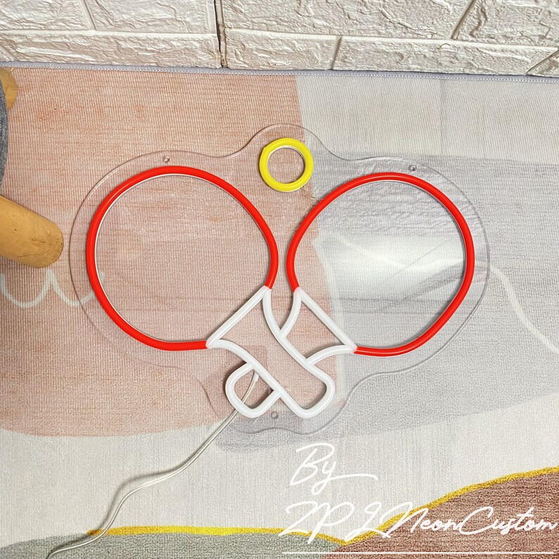 May include: A neon sign in the shape of a pair of ping pong paddles with red outlines and white handles. A yellow circle is above the paddles. The sign is on a clear acrylic backing. The text "By ZP Neon Custom" is at the bottom.