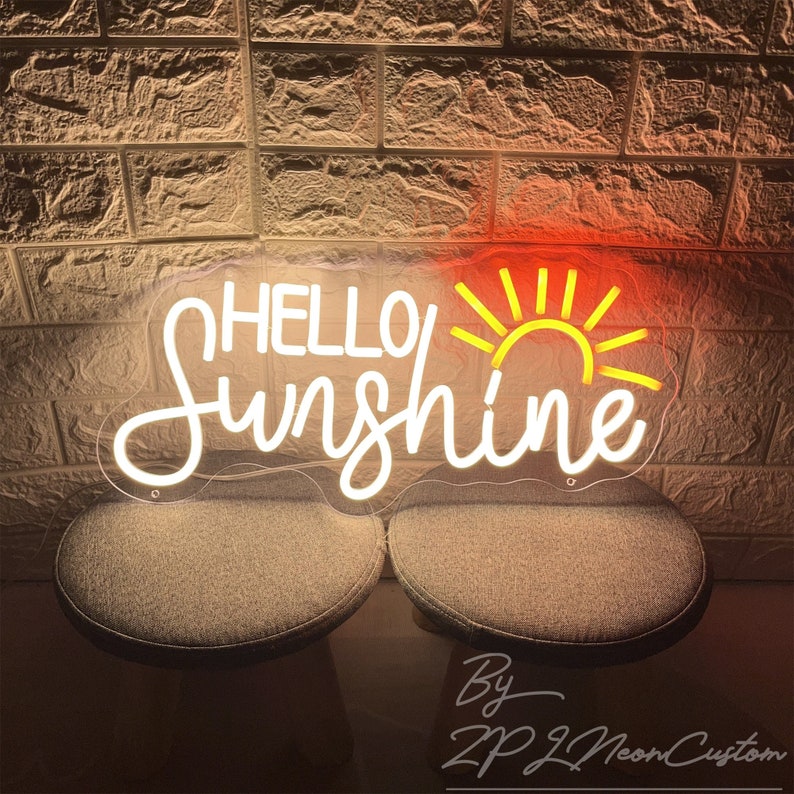 May include: A neon sign with the words "HELLO Sunshine" in white script and a yellow sun design. The sign is mounted on a clear backing and is illuminated against a brick wall.