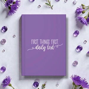 May include: A lilac-coloured journal with the text "FIRST THINGS FIRST. daily text" in white script. The journal is surrounded by purple flowers, clear and purple gems, and small clear beads on a white surface.