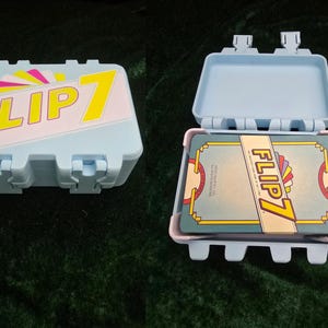 May include: A light blue plastic case with the text "FLIP 7" in yellow and pink. The case is shown closed and open, revealing a deck of cards with the same logo. The case has a rectangular shape with rounded corners.