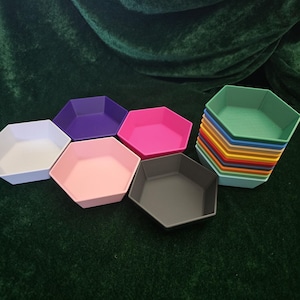 May include: A selection of hexagonal trays in a variety of colours, including white, purple, pink, grey, and green. A stack of similar trays in multiple colours is also visible. The trays are arranged on a dark green surface.