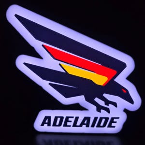 May include: A patch featuring a stylised black bird in flight with red and yellow wing accents. The design is outlined in a glowing white border against a dark background. Below the bird, the word ADELAIDE is printed in bold black capital letters.