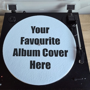 May include: Black turntable with a light blue record featuring the text "Your Favourite Album Cover Here" in black. The turntable has a clear plastic cover and is set on a wooden surface.