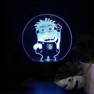 May include: A blue illuminated acrylic night light with a Minion character design. The light is circular, with the Minion etched into the surface, standing on a black base. The background is dark, with some green foliage visible.