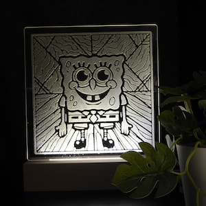 May include: An illuminated glass panel with an etched image of SpongeBob SquarePants. The cartoon character is smiling, with his arms at his sides. The panel is set on a wooden base, with a dark background and a potted plant.