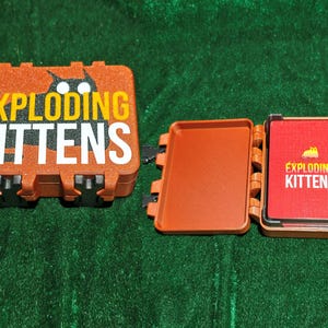 May include: A brown Exploding Kittens card game set. The game box is open, revealing a tray and a deck of red cards. The box and cards feature the game's logo and a cartoon cat illustration.