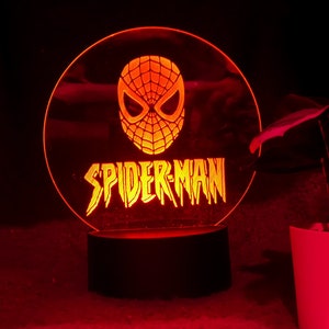 May include: A Spider-Man themed night light. The design features the Spider-Man mask above the word "SPIDER-MAN" in a stylized font. The light emits a warm orange glow against a dark background.