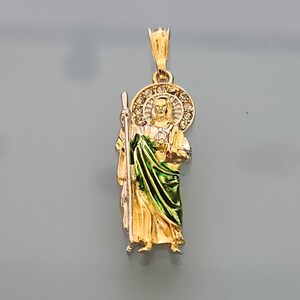 May include: Gold pendant featuring a detailed depiction of Saint Christopher, holding a staff and wearing a green robe. The pendant is adorned with a halo of clear stones.