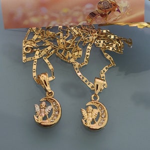 May include: Two gold necklaces with angel and crescent moon pendants. The necklaces are on a blue background. The text "Ashley Accesorios" and "Lo mas bello para Ti!" is visible on a card behind the necklaces.
