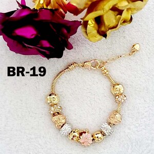 May include: Gold-toned bracelet with decorative charms, including floral and crystal designs. The bracelet is displayed with gold-colored roses and a deep pink rose. The text "BR-19" is visible.