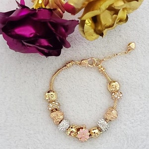 May include: A gold-toned bracelet with decorative charms, including floral and crystal-studded beads. The bracelet is displayed with gold-colored roses and a deep purple rose.