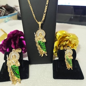 May include: A gold chain necklace with a pendant featuring a saint figure with green and white enamel. The pendant is surrounded by clear rhinestones. The necklace is displayed on a black stand. There are two smaller pendants with the same design on black stands.