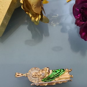 May include: Gold-tone pendant featuring a green and gold depiction of a saint holding a staff. The pendant is approximately 1 inch in length and is shown next to a ruler for size reference.