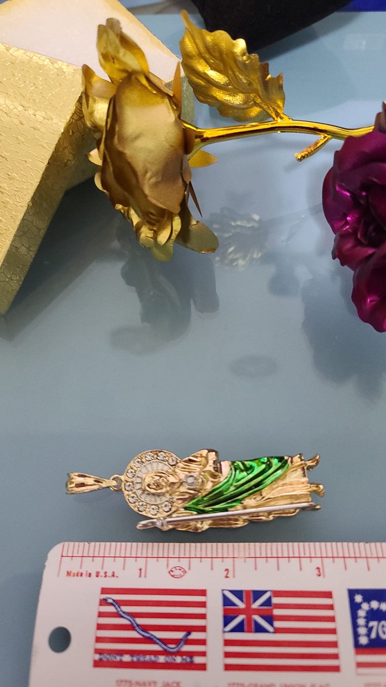 May include: Gold-tone pendant featuring a green and gold depiction of a saint holding a staff. The pendant is approximately 1 inch in length and is shown next to a ruler for size reference.