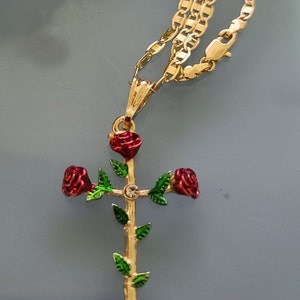 May include: A gold chain necklace with a pendant featuring a gold cross with three red roses and green leaves. The cross has a small clear rhinestone in the center.