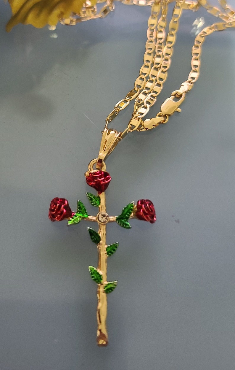 May include: A gold chain necklace with a pendant featuring a gold cross with three red roses and green leaves. The cross has a small clear rhinestone in the center.