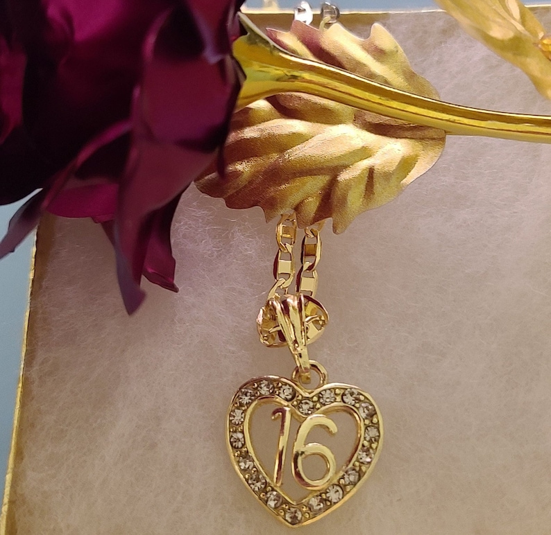 May include: A gold heart-shaped pendant necklace with clear rhinestones and the number 16 in the center. The necklace is hanging from a gold chain.