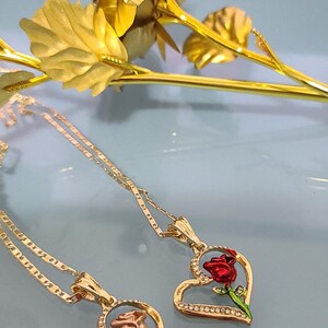 May include: Two gold necklaces with heart-shaped pendants featuring a rose design. One rose is red and the other is pink.