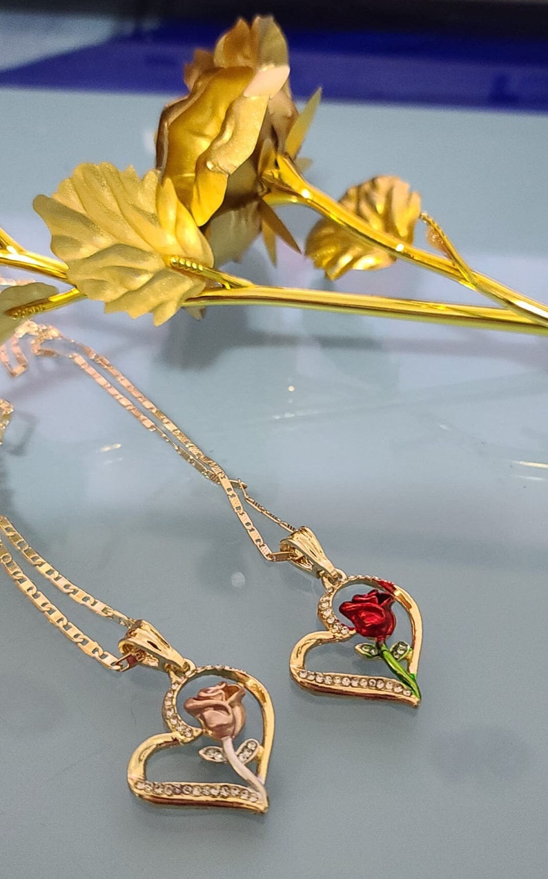 May include: Two gold necklaces with heart-shaped pendants featuring a rose design. One rose is red and the other is pink.