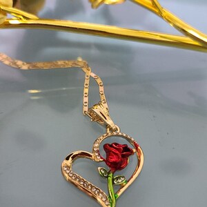 May include: A gold heart-shaped pendant necklace with a red rose and green leaves inside the heart. The pendant is adorned with small clear rhinestones.