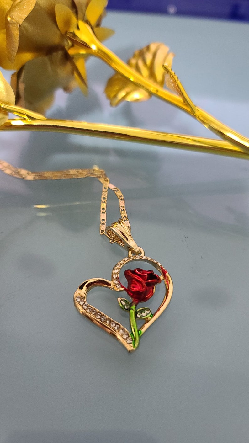 May include: A gold heart-shaped pendant necklace with a red rose and green leaves inside the heart. The pendant is adorned with small clear rhinestones.