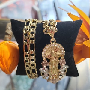 May include: A gold chain necklace with a pendant featuring a religious figure, likely the Virgin Mary, surrounded by roses. The chain is a Cuban link style and is displayed on a black velvet surface.