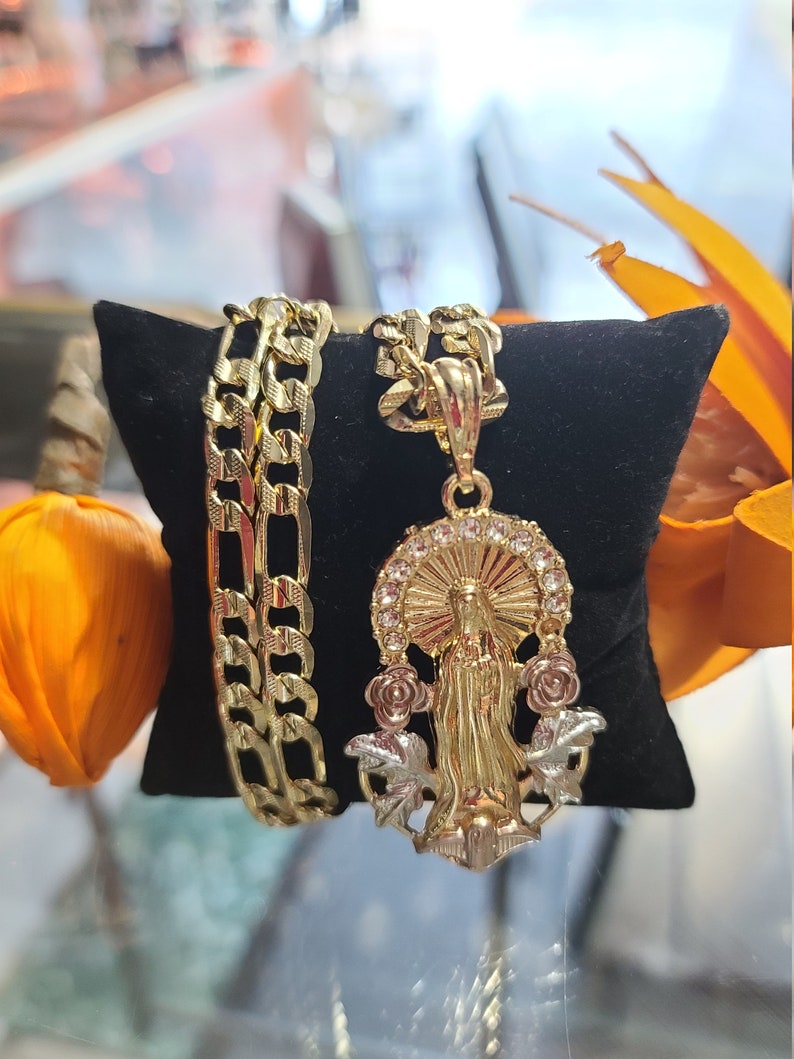 May include: A gold chain necklace with a pendant featuring a religious figure, likely the Virgin Mary, surrounded by roses. The chain is a Cuban link style and is displayed on a black velvet surface.