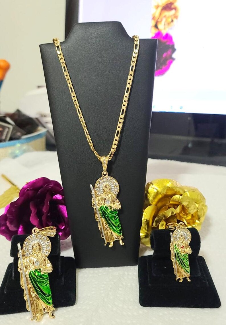 May include: A gold chain necklace with a pendant featuring a saint figure with green and white enamel. The pendant is surrounded by clear rhinestones. The necklace is displayed on a black stand. There are two smaller pendants with the same design on black stands.