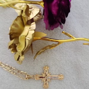 K&ouml;nnte beinhalten: Eine goldfarbene Halskette mit einem Kreuzanh&auml;nger, neben zwei goldfarbenen Rosen mit lila und goldenen Bl&uuml;tenbl&auml;ttern. Der Kreuzanh&auml;nger hat aufwendige Details. Das Ensemble liegt auf einem weichen, wei&szlig;en Hintergrund.