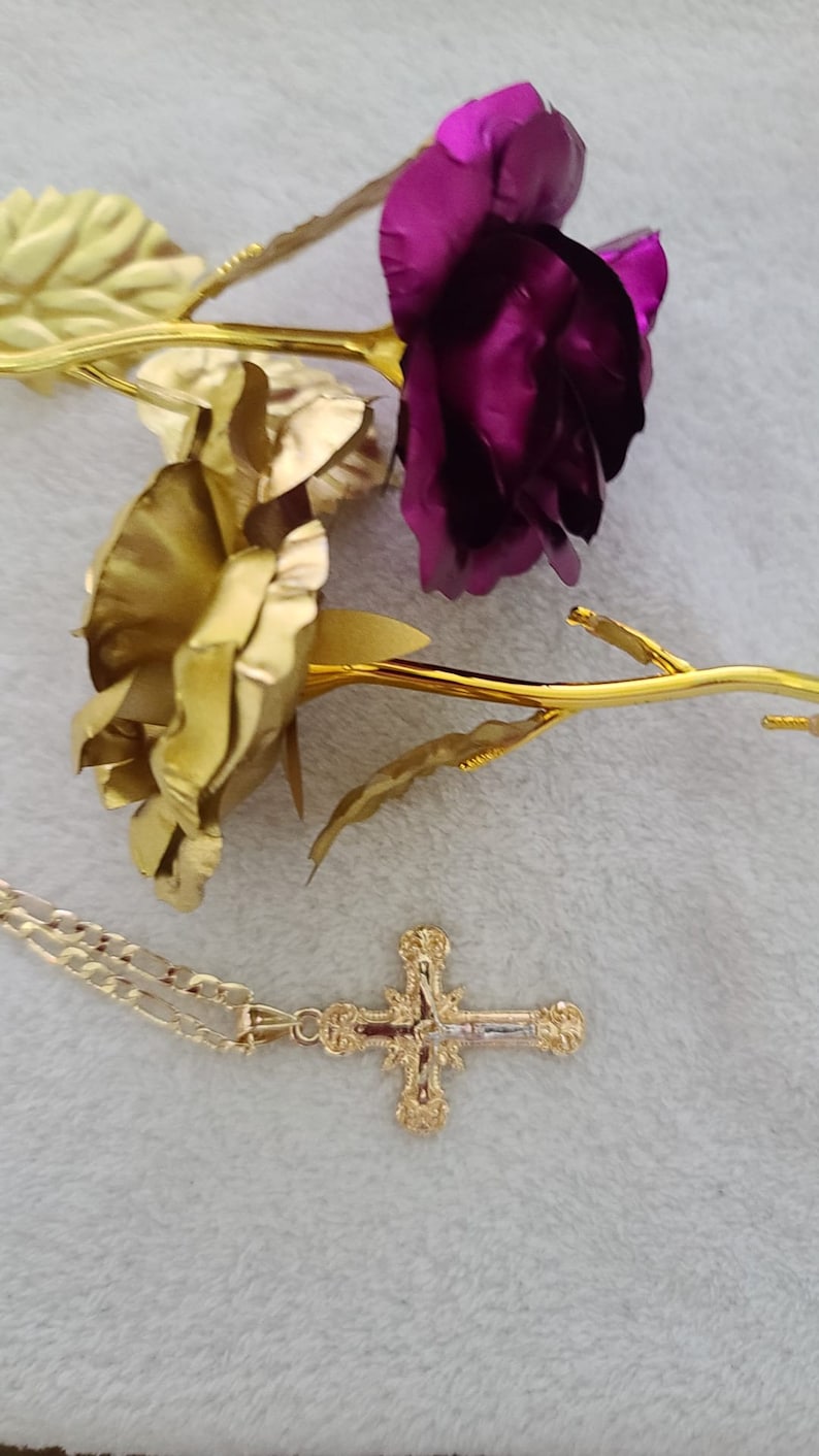 K&ouml;nnte beinhalten: Eine goldfarbene Halskette mit einem Kreuzanh&auml;nger, neben zwei goldfarbenen Rosen mit lila und goldenen Bl&uuml;tenbl&auml;ttern. Der Kreuzanh&auml;nger hat aufwendige Details. Das Ensemble liegt auf einem weichen, wei&szlig;en Hintergrund.