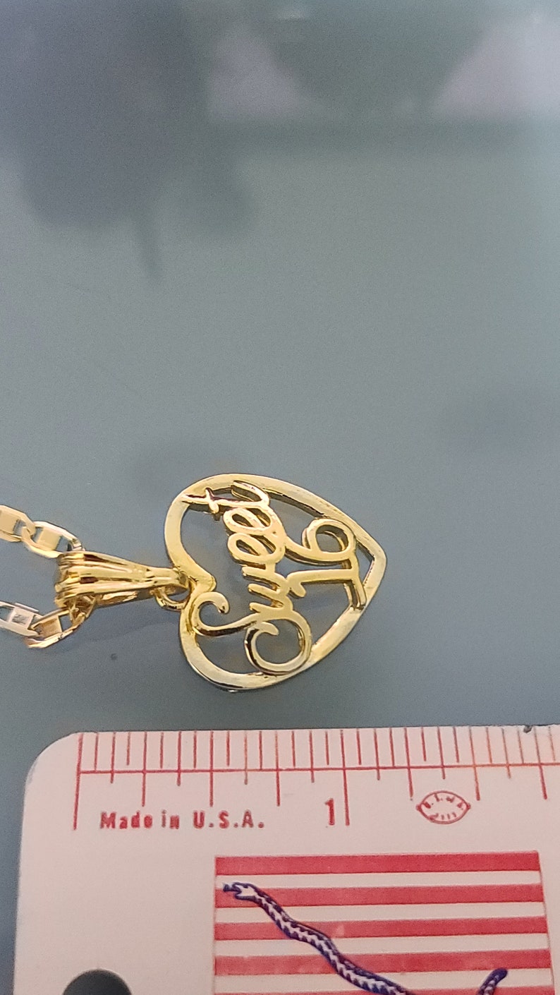May include: Gold heart-shaped pendant necklace with the word "Sweet" inscribed inside. The necklace is on a gold chain and is next to a ruler.