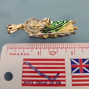 May include: A gold-colored pendant depicting a religious figure with a green and gold robe. The pendant is approximately 1 inch in length and is measured against a ruler with a "Don't Tread on Me" flag design.