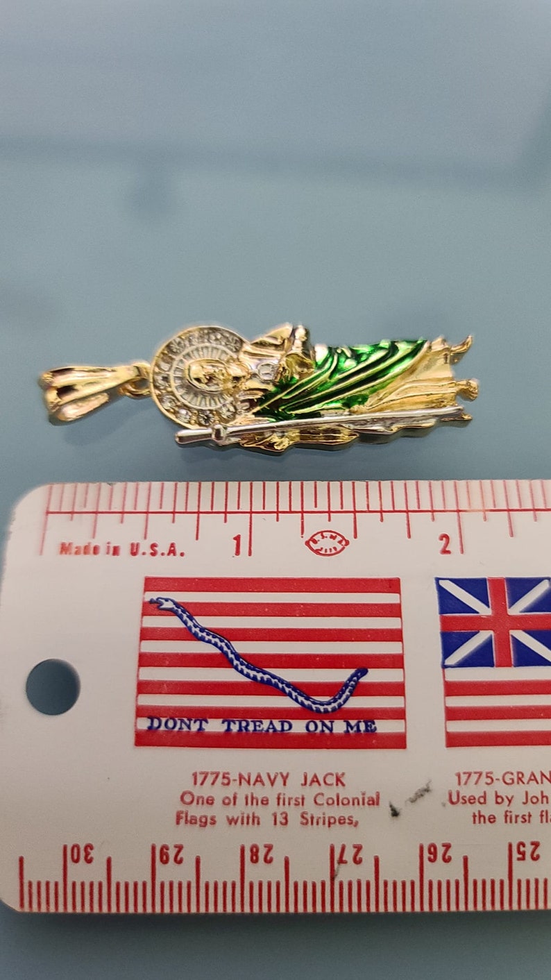 May include: A gold-colored pendant depicting a religious figure with a green and gold robe. The pendant is approximately 1 inch in length and is measured against a ruler with a "Don't Tread on Me" flag design.