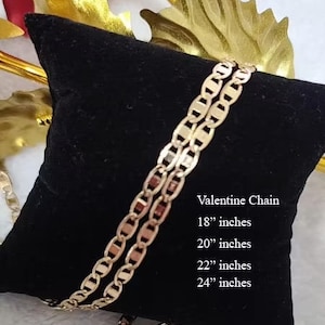 May include: A gold-colored chain necklace on a black velvet cushion. The chain is a mariner style with oval links. The text "Valentine Chain" is printed on the cushion, along with the available sizes: 18", 20", 22", and 24" inches.