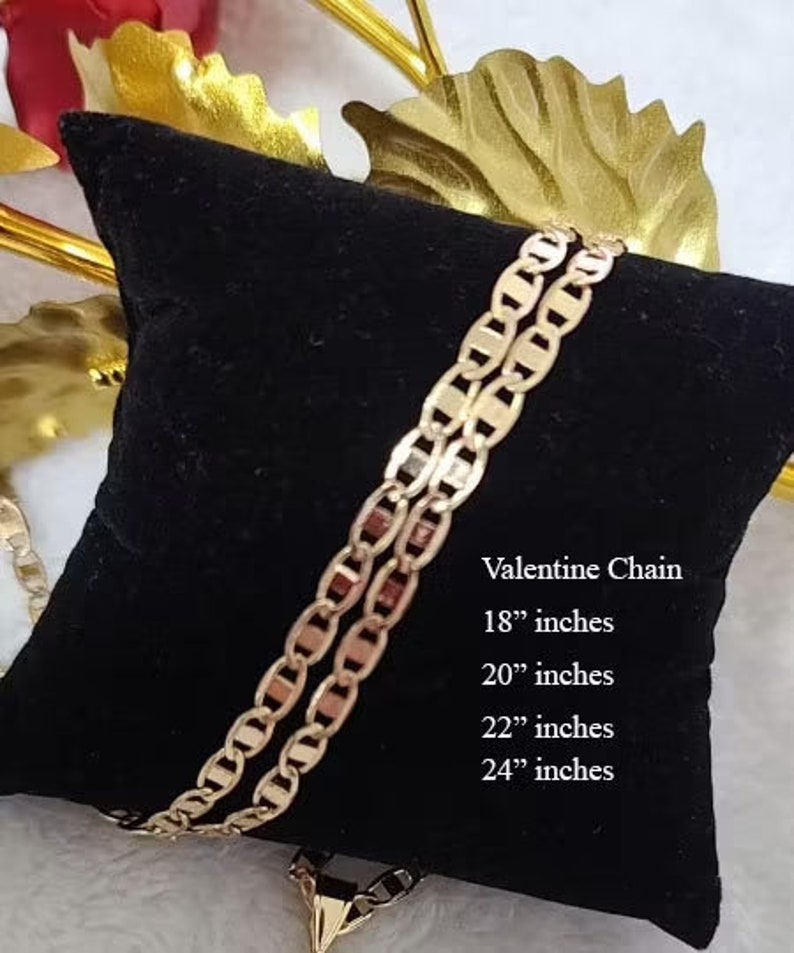 May include: A gold-colored chain necklace on a black velvet cushion. The chain is a mariner style with oval links. The text "Valentine Chain" is printed on the cushion, along with the available sizes: 18", 20", 22", and 24" inches.