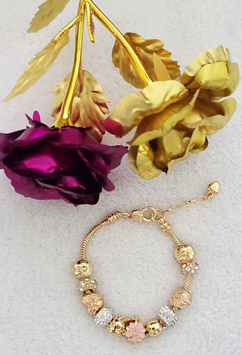 May include: A gold-toned bracelet with decorative charms, including floral and crystal-studded beads. The bracelet is displayed with gold-colored roses and a deep purple rose.