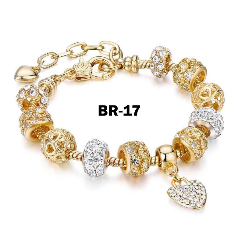 May include: Gold-toned bracelet with various decorative beads, including clear crystal-like accents and openwork designs. A heart-shaped charm with crystal embellishments hangs from the bracelet. The text "BR-17" is visible.