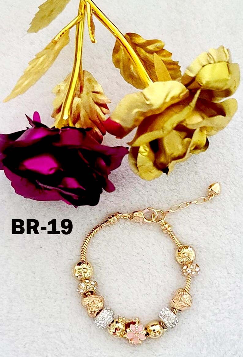 May include: Gold-toned bracelet with decorative charms, including floral and crystal designs. The bracelet is displayed with gold-colored roses and a deep pink rose. The text "BR-19" is visible.