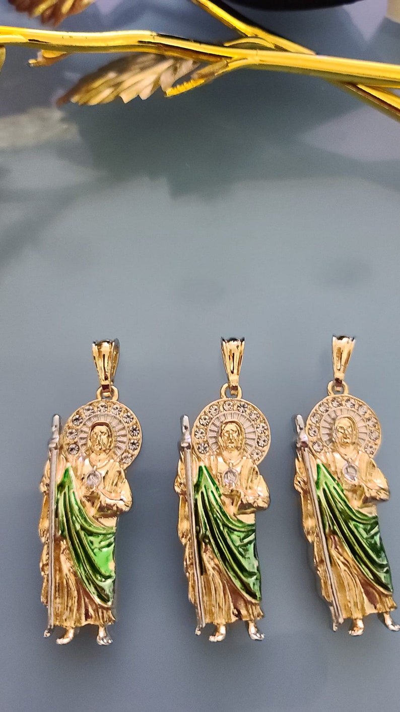 May include: Three gold pendants featuring a religious figure with a green robe and a halo of white stones. The pendants are on a light blue background.