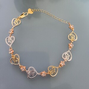May include: A gold and silver-toned bracelet featuring alternating heart-shaped charms with the number 15 inside and small round clear stones. The bracelet includes a delicate chain link section with a heart-shaped clasp at the end.
