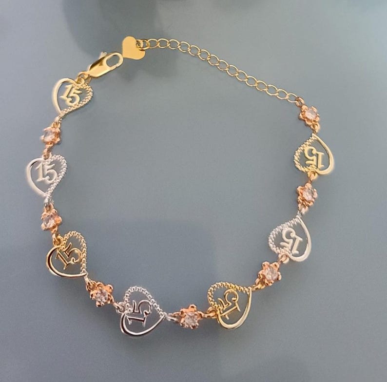 May include: A gold and silver-toned bracelet featuring alternating heart-shaped charms with the number 15 inside and small round clear stones. The bracelet includes a delicate chain link section with a heart-shaped clasp at the end.