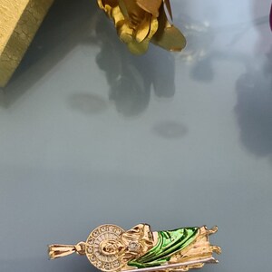 May include: Gold-tone pendant featuring a green and gold depiction of a religious figure holding a staff. The pendant is approximately 2 inches long and is measured against a ruler. "Made in U.S.A." is printed on the ruler.