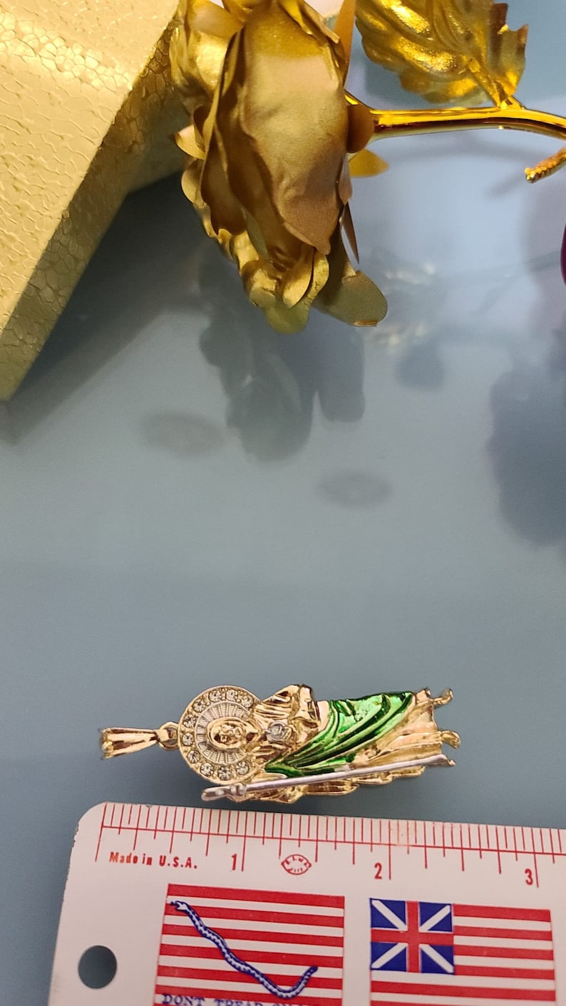 May include: Gold-tone pendant featuring a green and gold depiction of a religious figure holding a staff. The pendant is approximately 2 inches long and is measured against a ruler. "Made in U.S.A." is printed on the ruler.