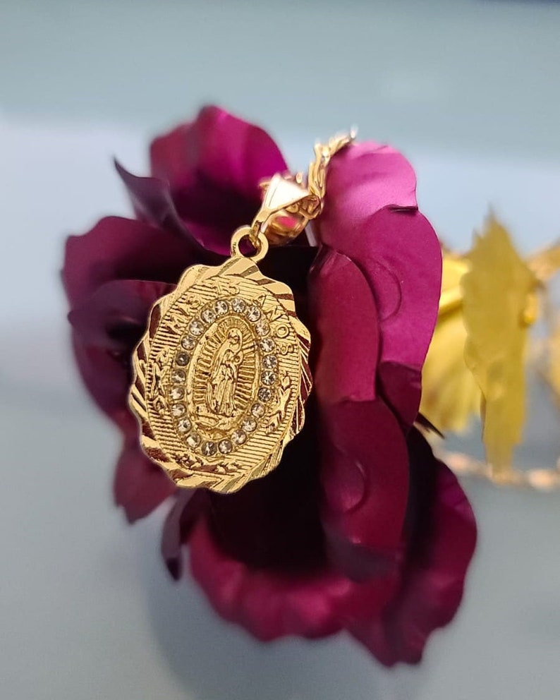 May include: Gold-tone pendant necklace featuring a detailed oval-shaped charm with a depiction of the Virgin Mary surrounded by sparkling rhinestones. The charm is attached to a delicate chain.