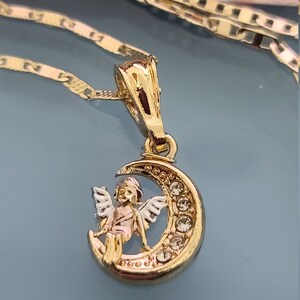 May include: A gold pendant necklace featuring a crescent moon with a small angel sitting on it. The moon is decorated with clear rhinestones.