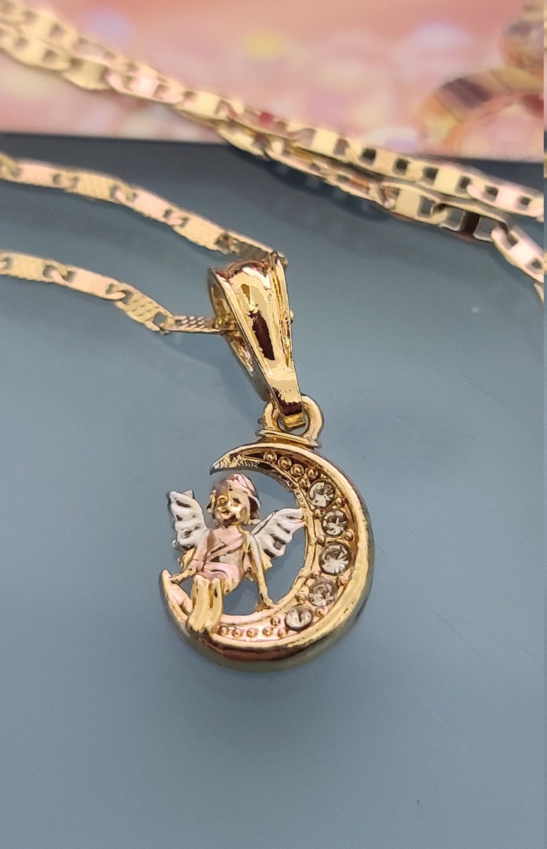 May include: A gold pendant necklace featuring a crescent moon with a small angel sitting on it. The moon is decorated with clear rhinestones.