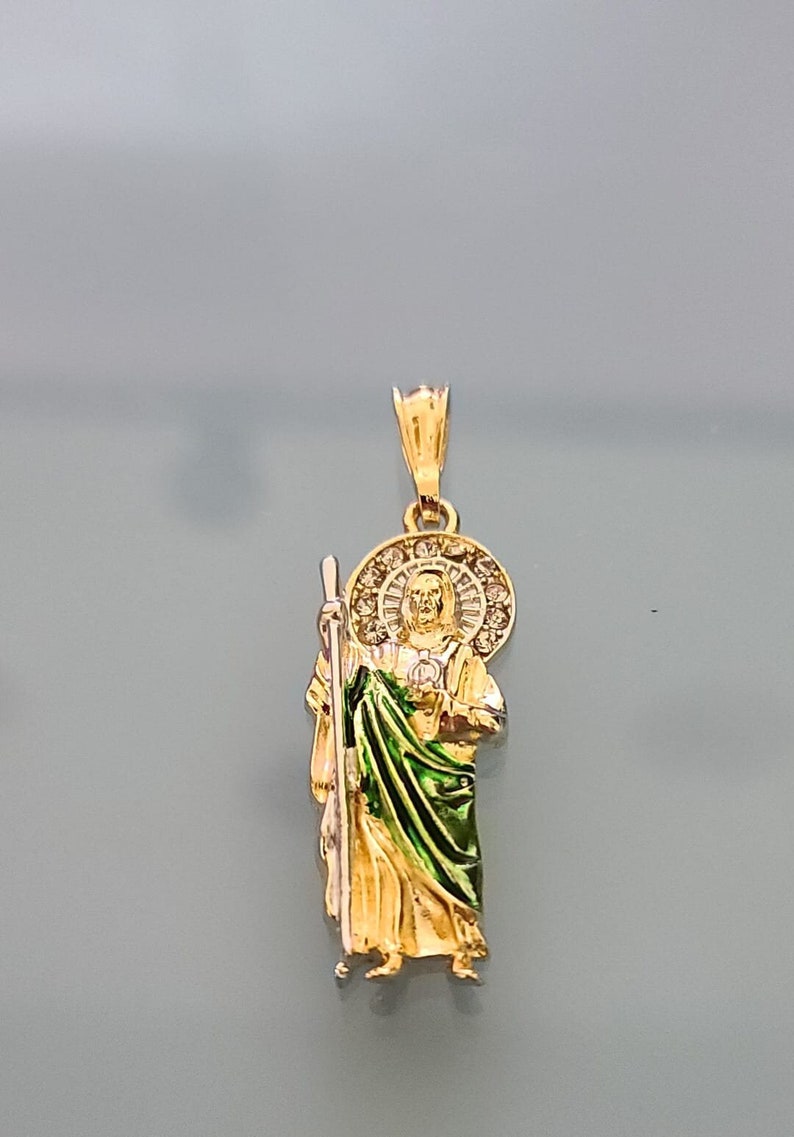 May include: Gold pendant featuring a detailed depiction of Saint Christopher, holding a staff and wearing a green robe. The pendant is adorned with a halo of clear stones.