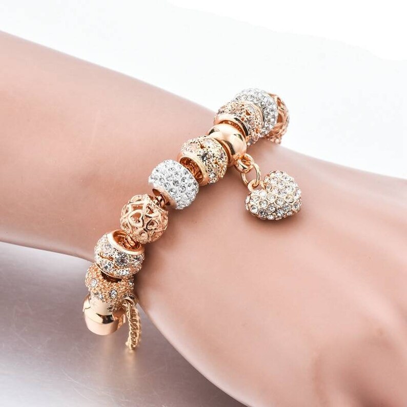 May include: Gold-toned charm bracelet with various decorative beads. The bracelet features beads with clear crystal accents, openwork designs, and a heart-shaped charm. The bracelet is designed to be worn on the wrist.
